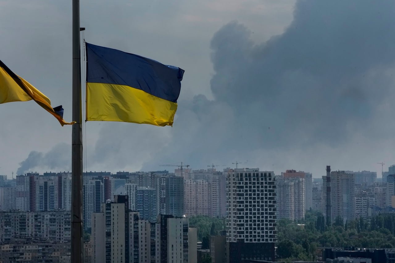 Ukrainian flags seen in foreground of image, with smoke rising above buildings in Kyiv, in the background.