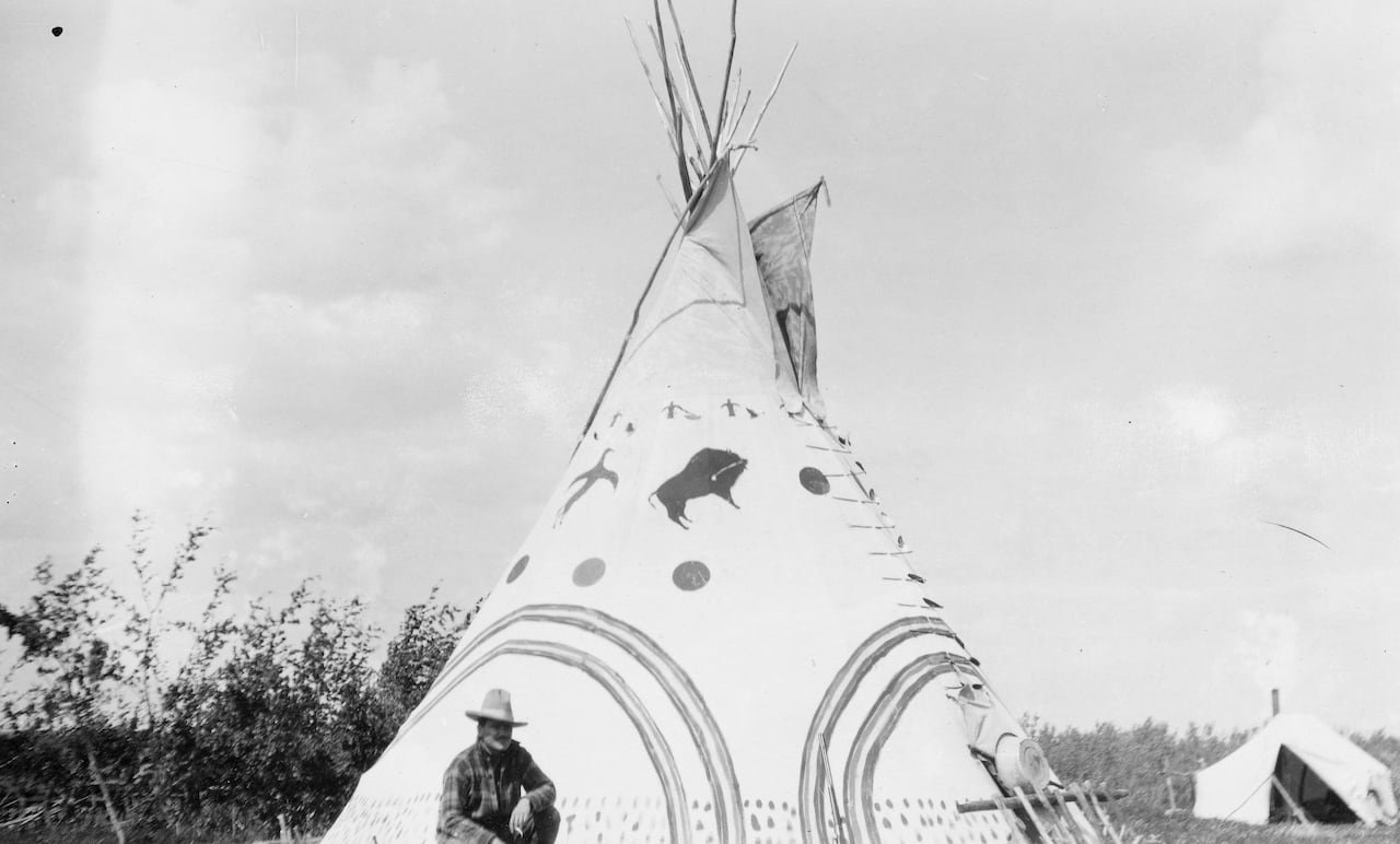 A black and white photograph of a teepee with a painted cover. A man wearing a Western hat kneels in front of it.