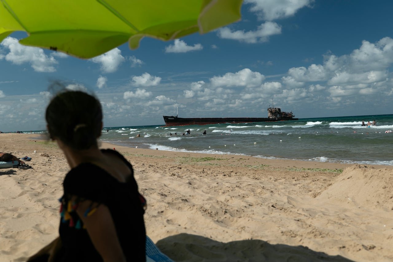 A woman on a beach looking out at the choppy waters and a giant cargo ship with visible damage near the shore.